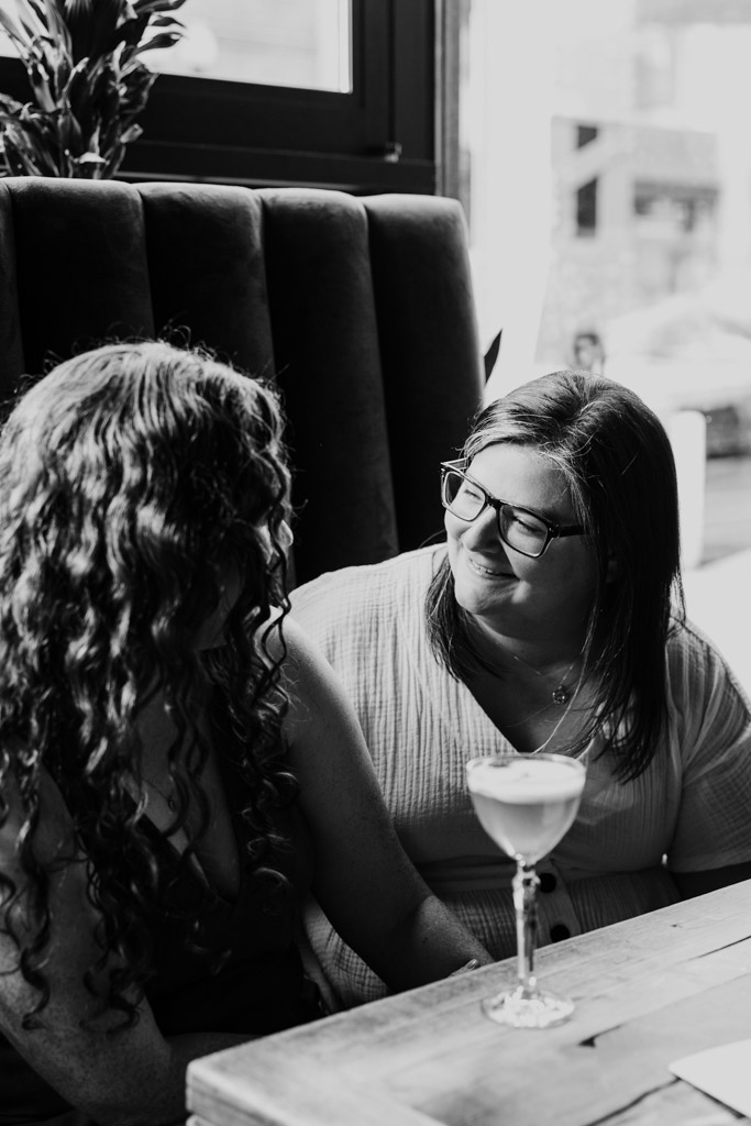Black and white photo of two women sitting at a table inside Clara cocktail bar in Wicker Park, smiling at each other with a cocktail glass in front of them