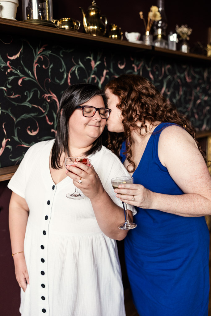 Two women smile, holding drinks as one kisses the other's cheek in Clara cocktail bar during their Wicker Park engagement session