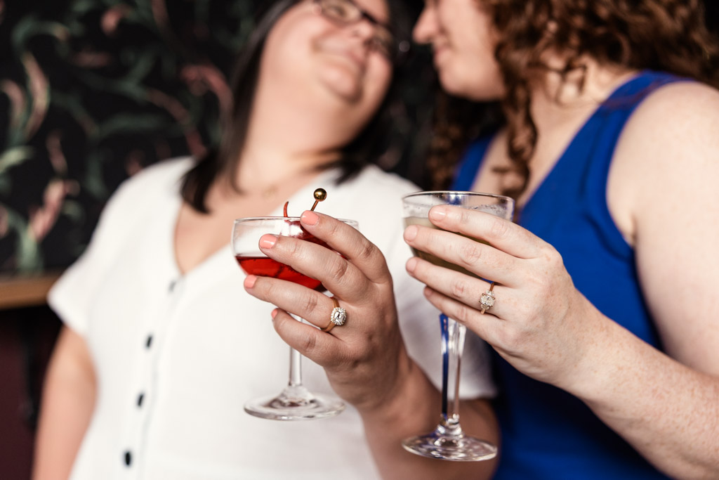Two women, both wearing rings, smiling and holding cocktails in their hands, inside Clara cocktail bar during their Wicker Park summer engagement session
