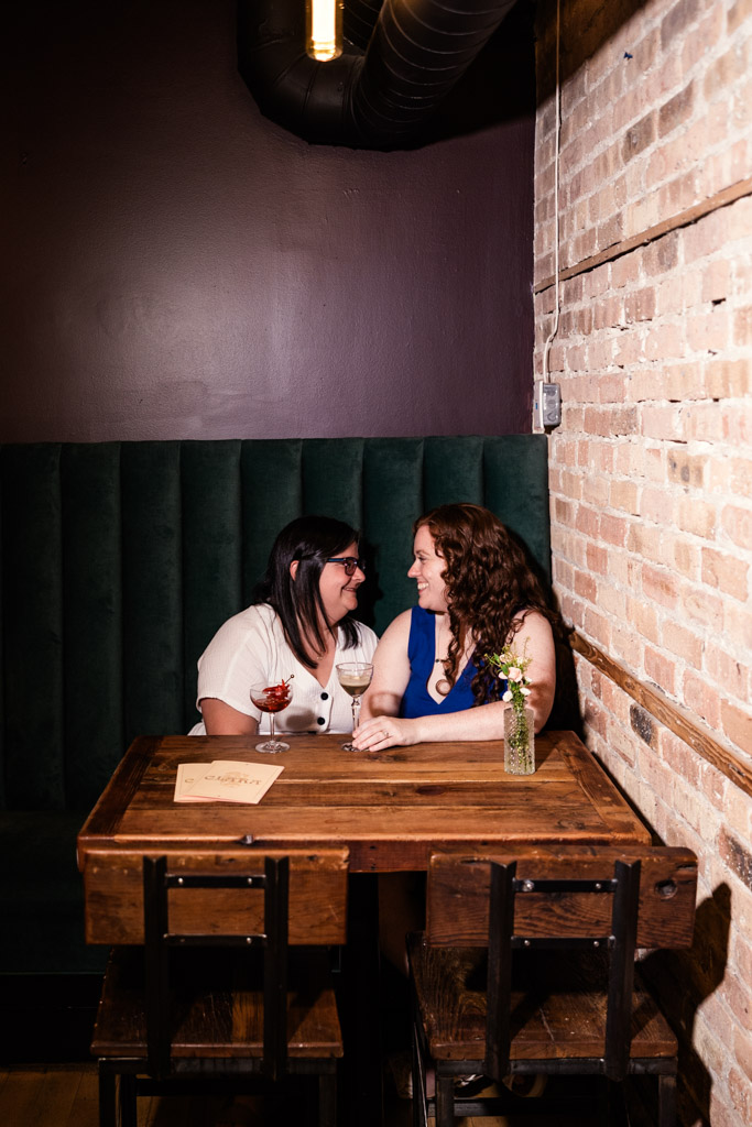 Two women smile at each other, sitting at a wooden table with drinks inside Clara cocktail bar during their Wicker Park engagement session
