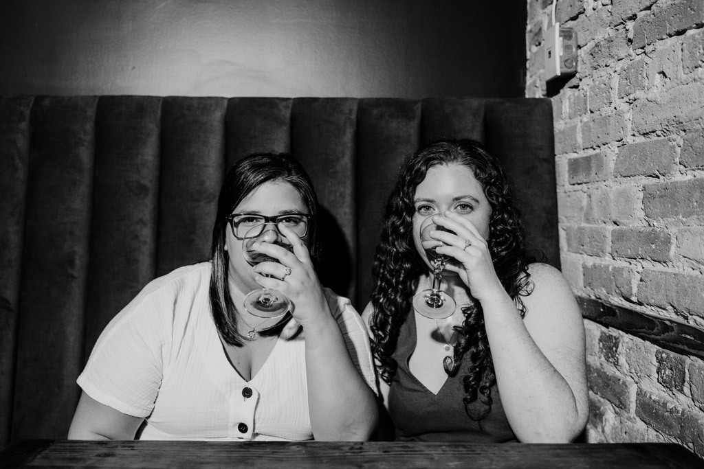 Black and white photo of two women sit ting side by side at a table, holding drinks and looking at the camera inside Clara cocktail bar during their Wicker Park engagement session