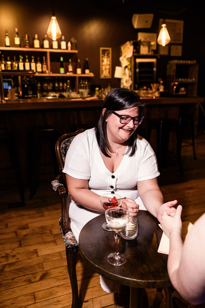 Woman in white dress smiling, sitting at a small table with her partner in the dimly lit Clara cocktail bar