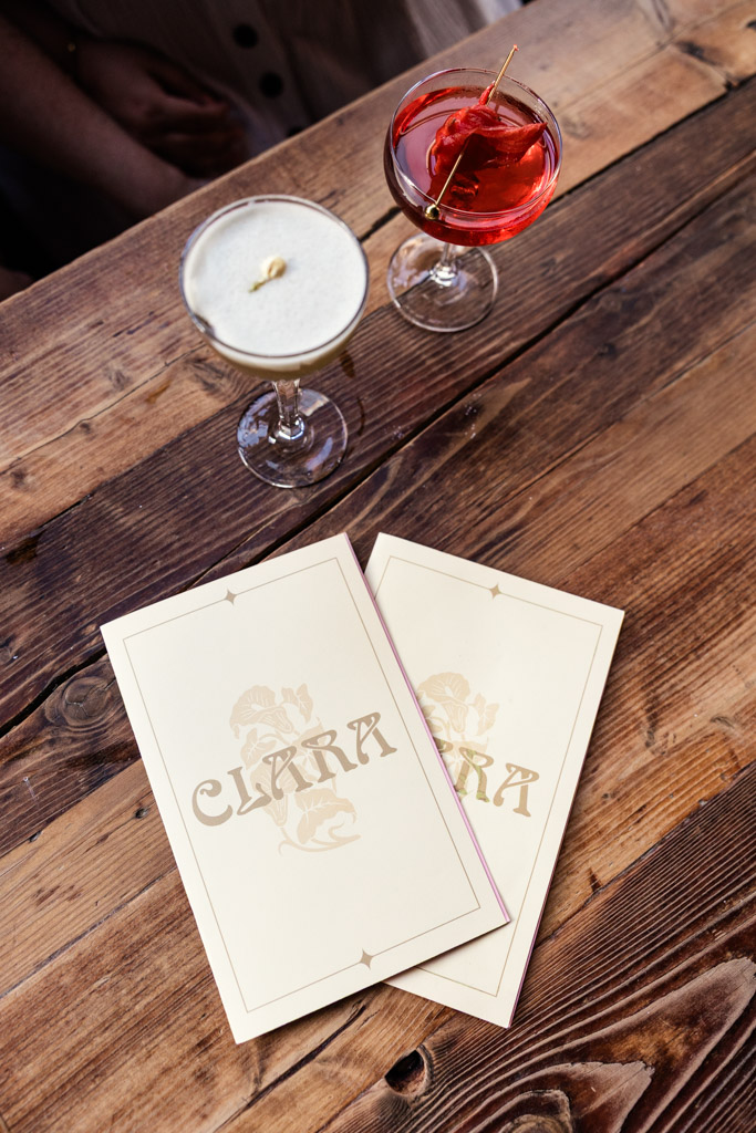 Two cocktails and two menus labeled rest on a rustic wooden table inside Clara cocktail bar