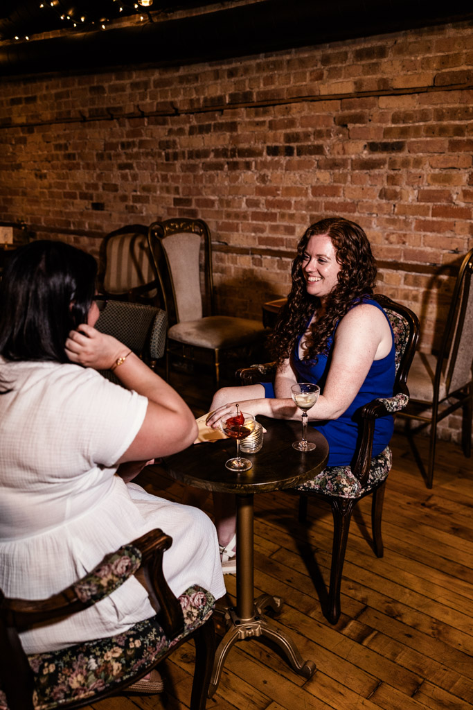 Two women sit at a small table in a cozy room, talking and enjoying drinks together during their Wicker Park engagement session