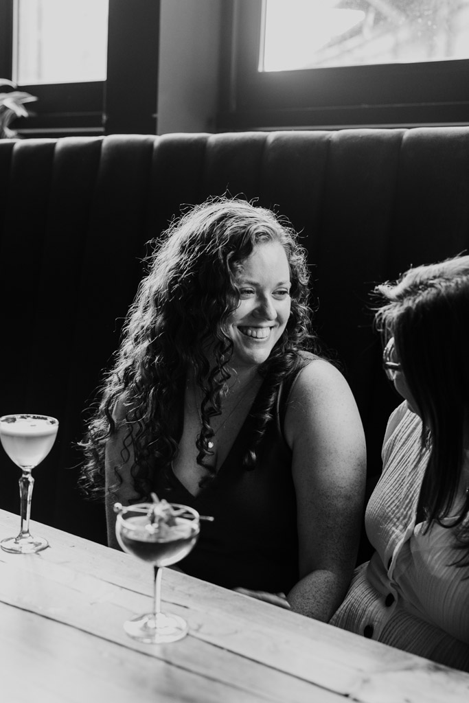 Black and white photo of two women sitting together at a table with cocktails in Clara cocktail bar in Wicker Park, laughing and enjoying each other's company