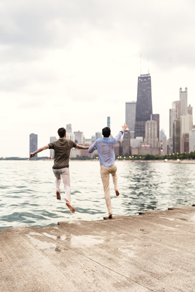 Engaged couple holding hands and joyfully jumping off a lakeside pier at North Avenue Beach with the Chicago skyline in the background