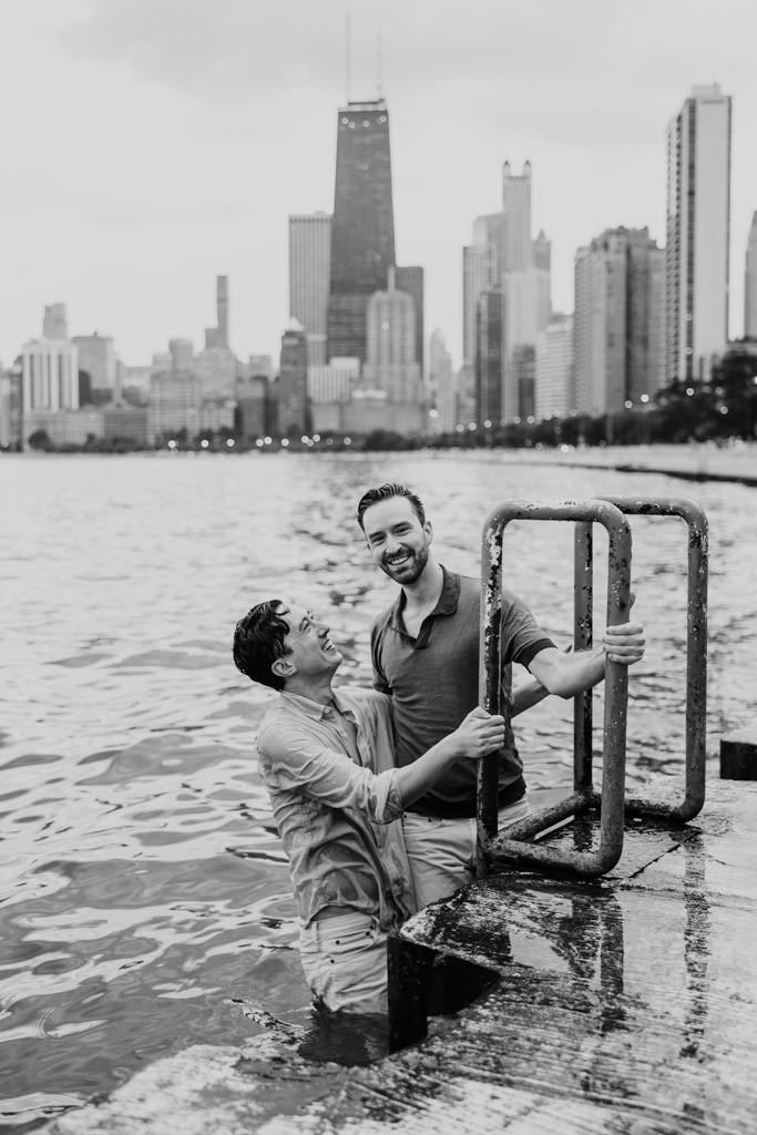 Black and white photo of two men smiling as they climb out of Lake Michigan on a metal ladder during their Chicago lakefront engagement session