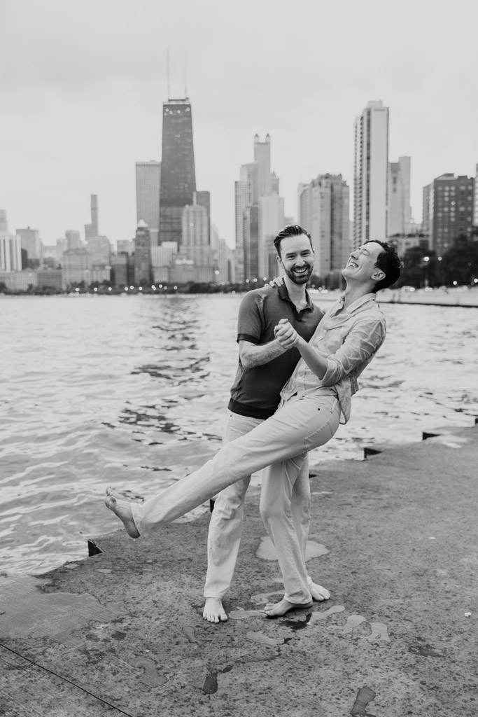 Two people laugh and dance barefoot by the water along the Chicago lakefront during their engagement session, with the city skyline in the background