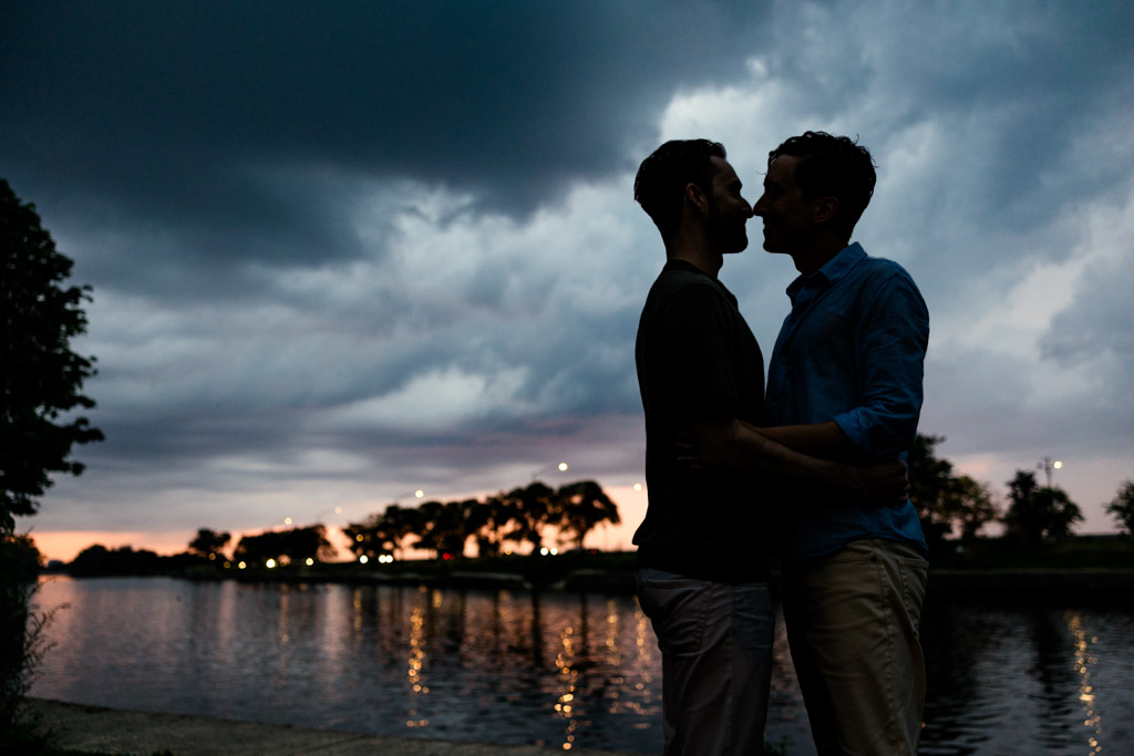 Two people embrace by a Lincoln Park pond at sunset, silhouetted against a dramatic cloudy sky
