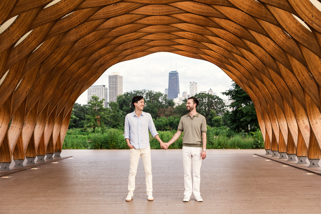 Two men hold hands under the Lincoln Park Honeycomb with the Chicago skyline visible in the background.