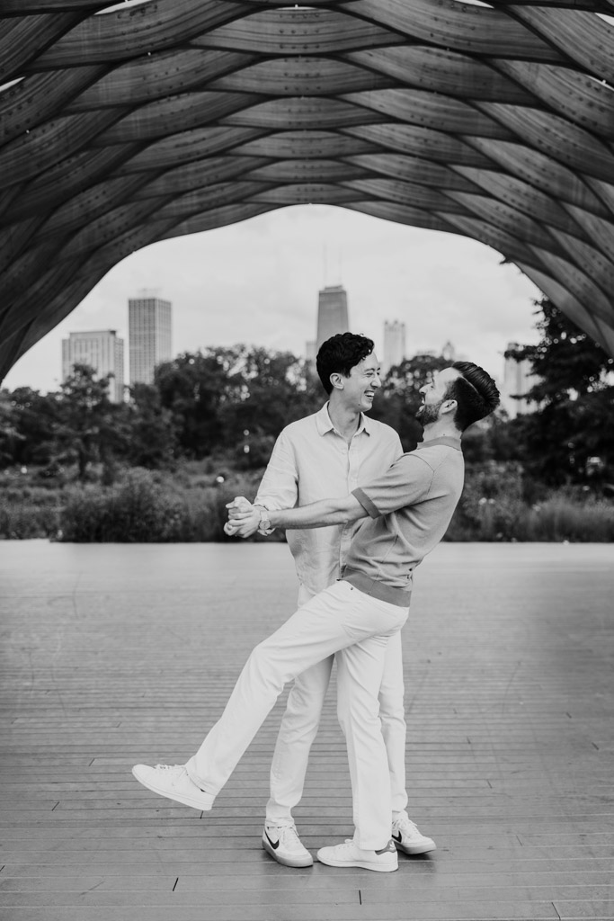Black and white photo of two men dancing under the Lincoln Park Honeycomb with skyscrapers in the background during their Chicago lakefront engagement session