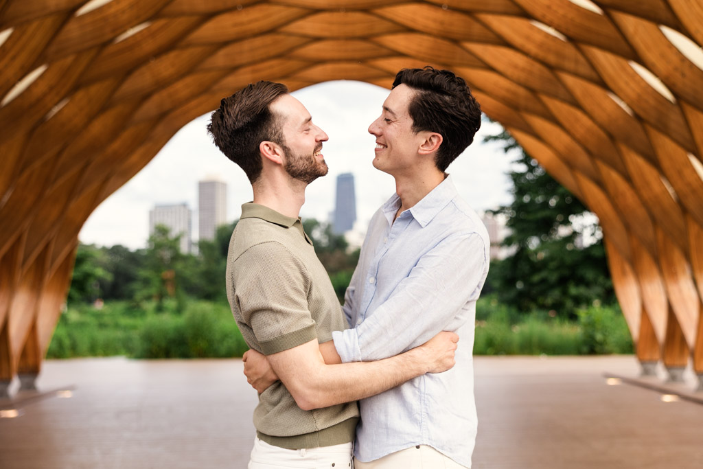 Two men smile and embrace under the Lincoln Park Honeycomb, with the Chicago skyline and lush greenery in the background