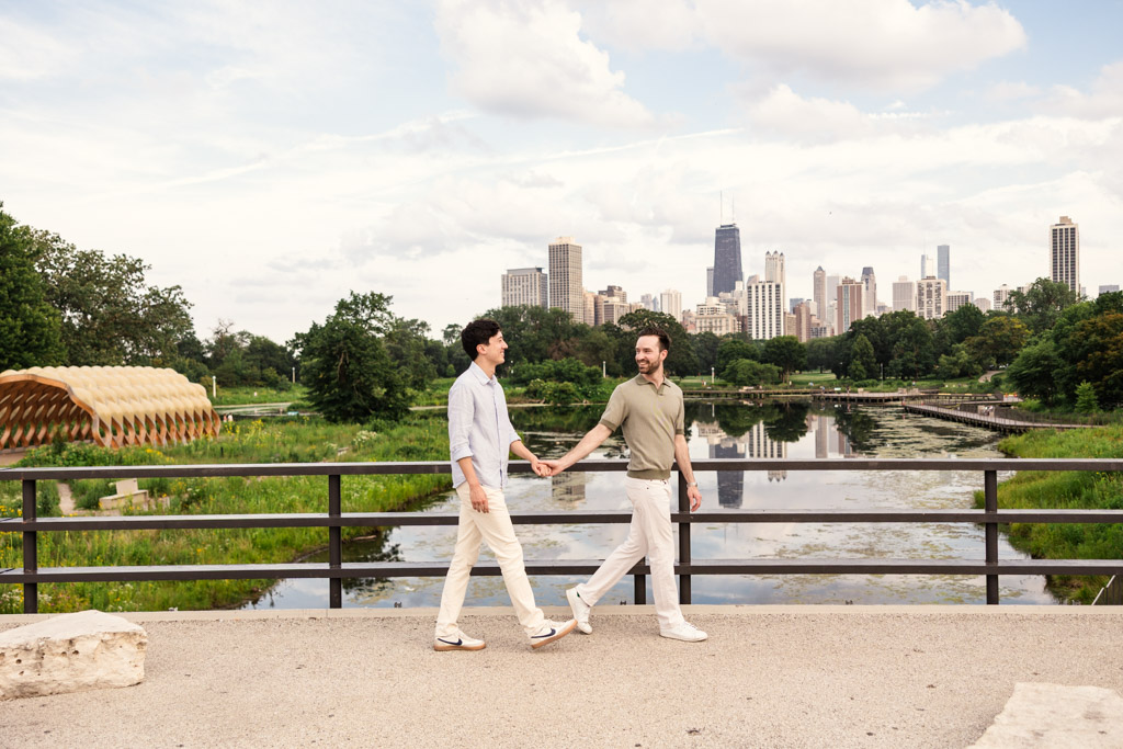 Two men holding hands and walking on a bridge in Lincoln Park, with downtown Chicago in the background