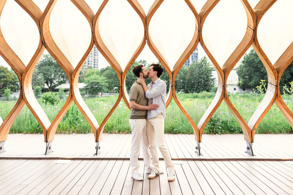 Two men kiss under the Lincoln Park Honeycomb with greenery and buildings in the background during their Chicago lakefront engagement session