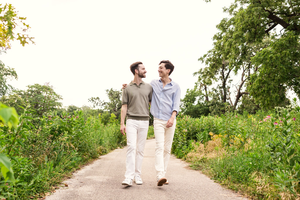 Two men walking on a path through greenery in Lincoln Park smiling at each other under a bright sky during their Chicago lakefront engagement session