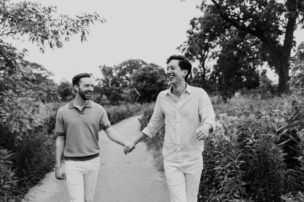 Black and white photo of engaged couple holding hands and laughing while walking on a Lincoln Park path surrounded by trees and greenery, during their Chicago lakefront engagement session