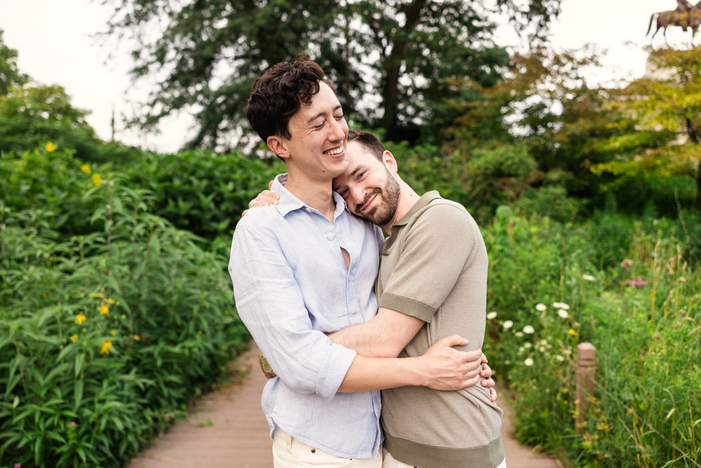 Two men stand on a Lincoln Park path, hugging and smiling, surrounded by green plants and trees during their Chicago lakefront engagement session