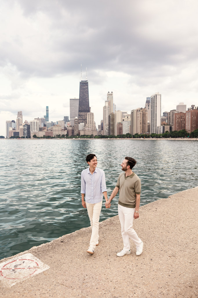 Two men hold hands and walk by the water on North Avenue Beach, with Chicago skyline and cloudy sky in the background during their lakefront engagement session