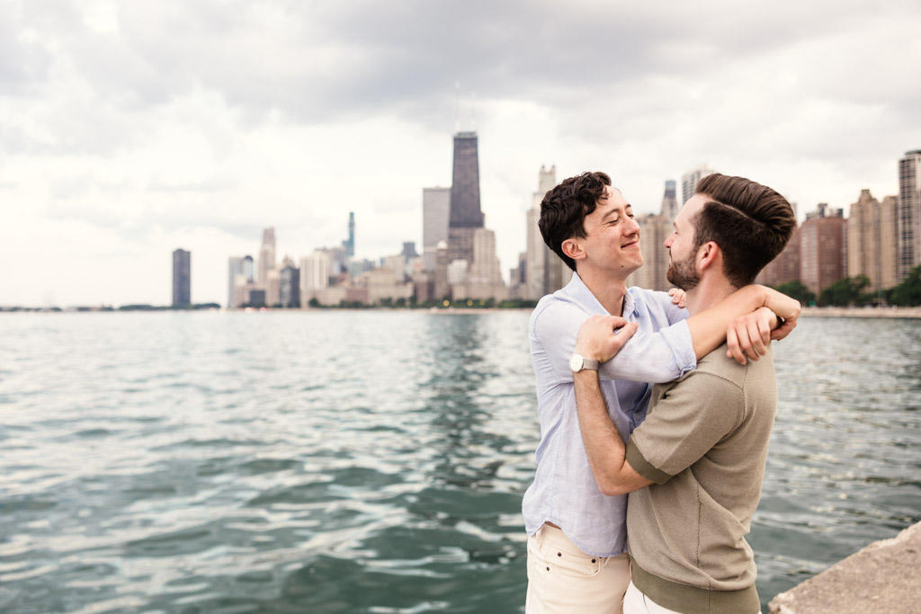 Two men embrace and smile by the lakefront at North Avenue Beach with Chicago skyline in the background on a cloudy day