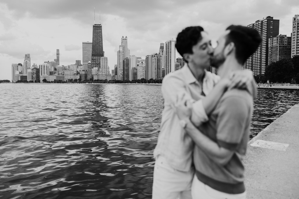 Creative black and white photo of two men kissing by the water along the Chicago lakefront, with focus on the city skyline and tall buildings in the background