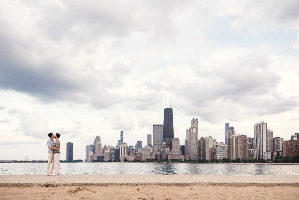 Engaged couple kisses on North Avenue beach along the Chicago lakefront, with city skyline and cloudy sky in the background