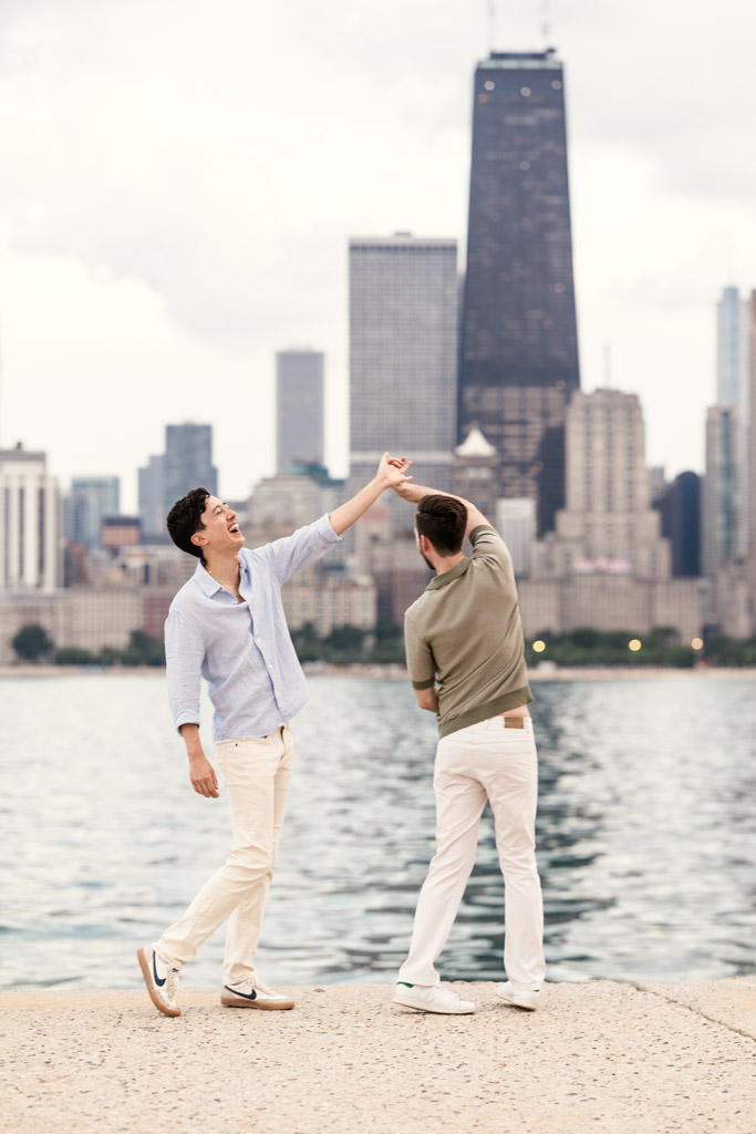 Two men dance together by the water along North Avenue Beach, with city skyline and tall buildings in the background during their Chicago engagement session