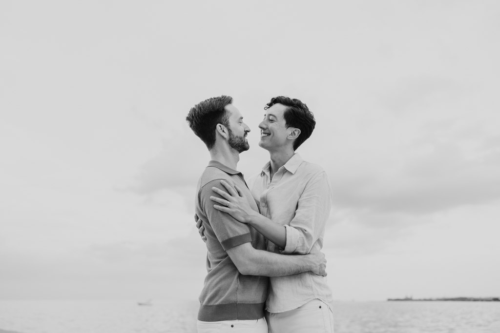 Black and white photo of couple smiling and embracing by Lake Michigan under a cloudy sky during their Chicago lakefront engagement session