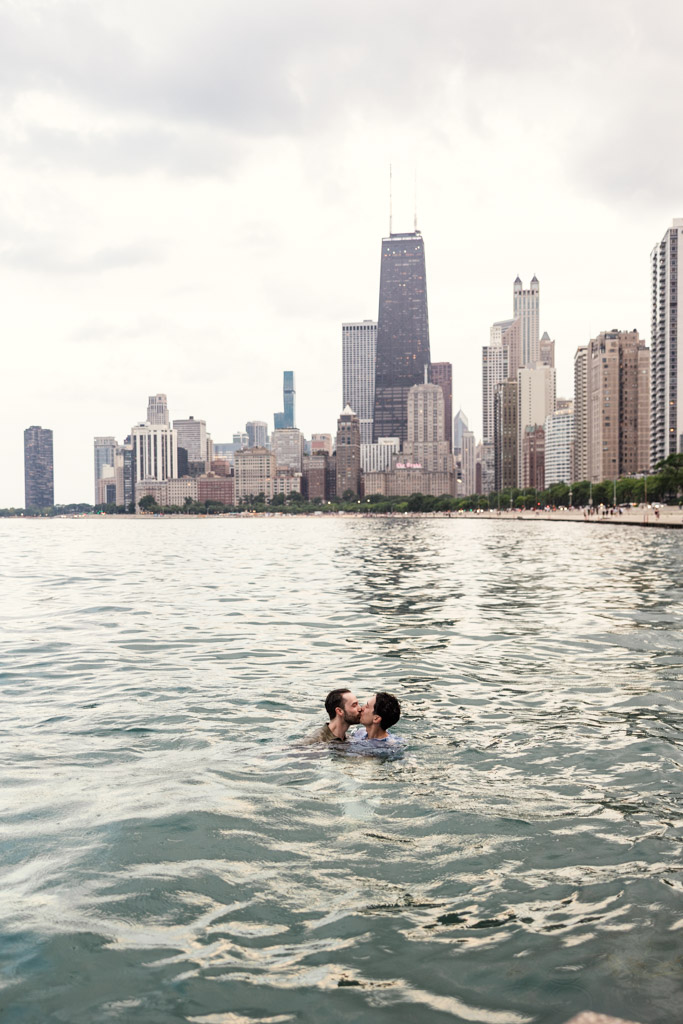 Two people kiss after jumping into Lake Michigan with Chicago skyline and tall buildings in the background under a cloudy sky during their Chicago lakefront engagement session