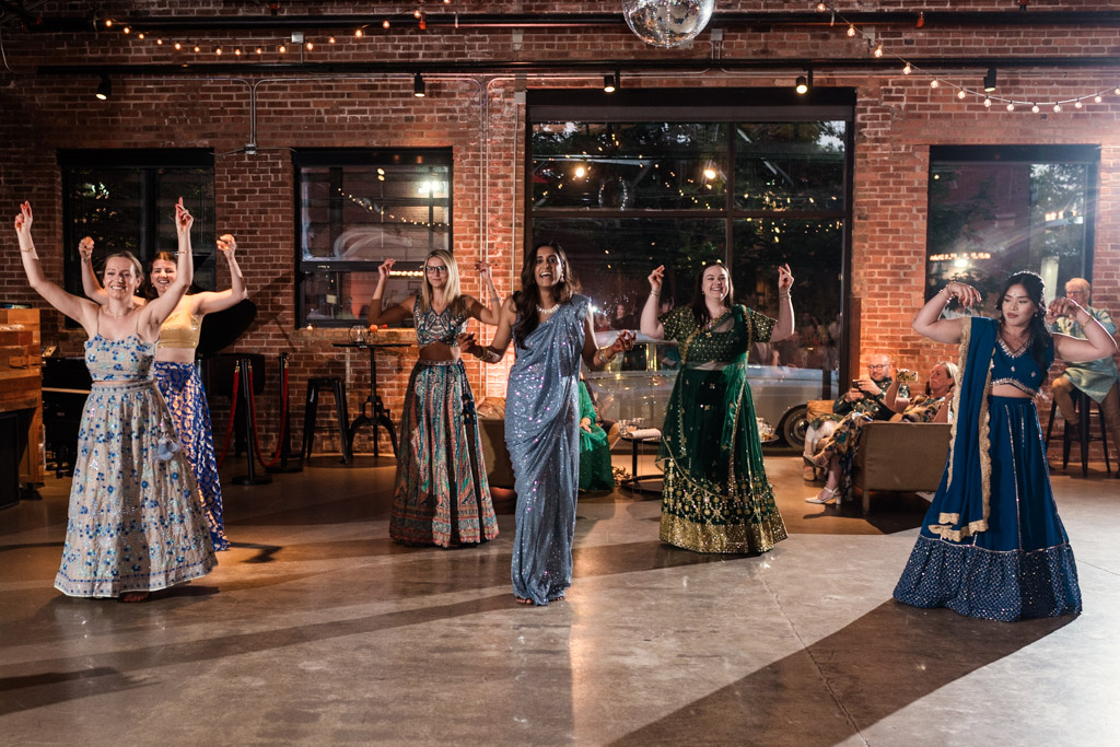 Six women in colorful dresses dance indoors under string lights, with people seated in the background during sangeet at Ovation Chicago