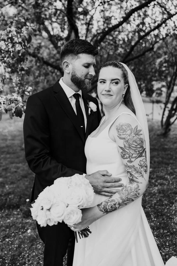 Black and white portrait of bride with tattoos holding flowers, groom in suit embraces her outdoors under trees in Horner Park