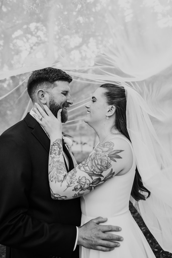 Black and white photo of bride and groom smiling at each other under a veil