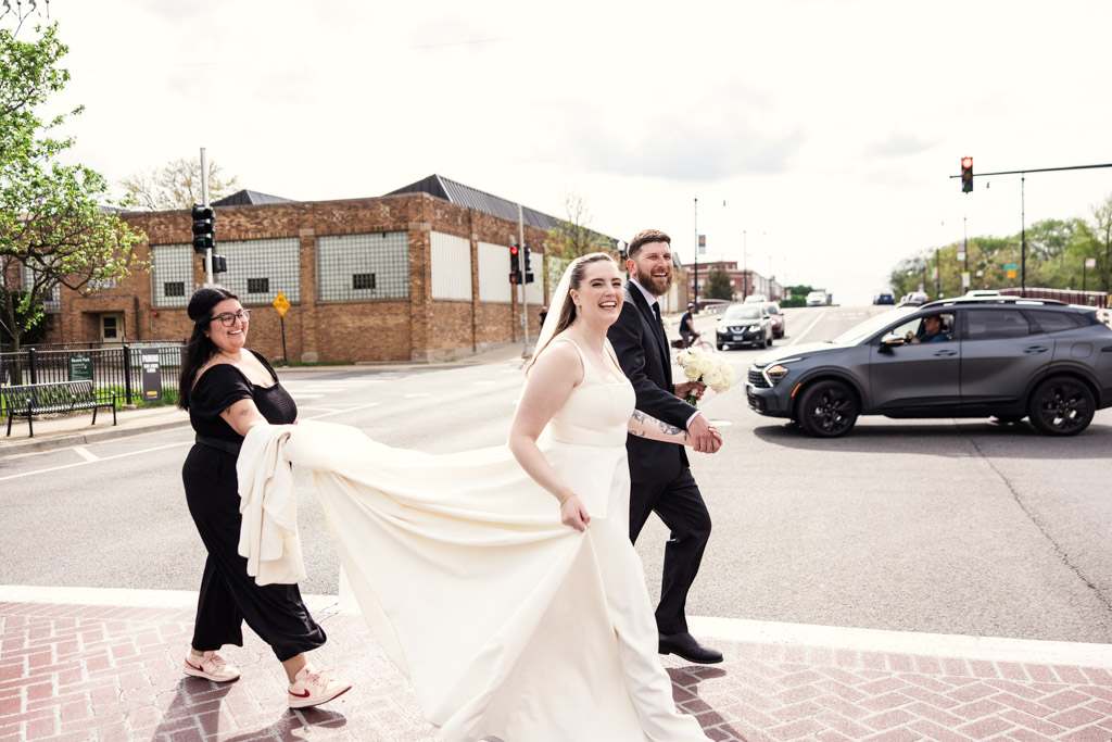 Bride, groom, and another woman cross street smiling near River Room; the woman holds bride's dress; cars and buildings in background