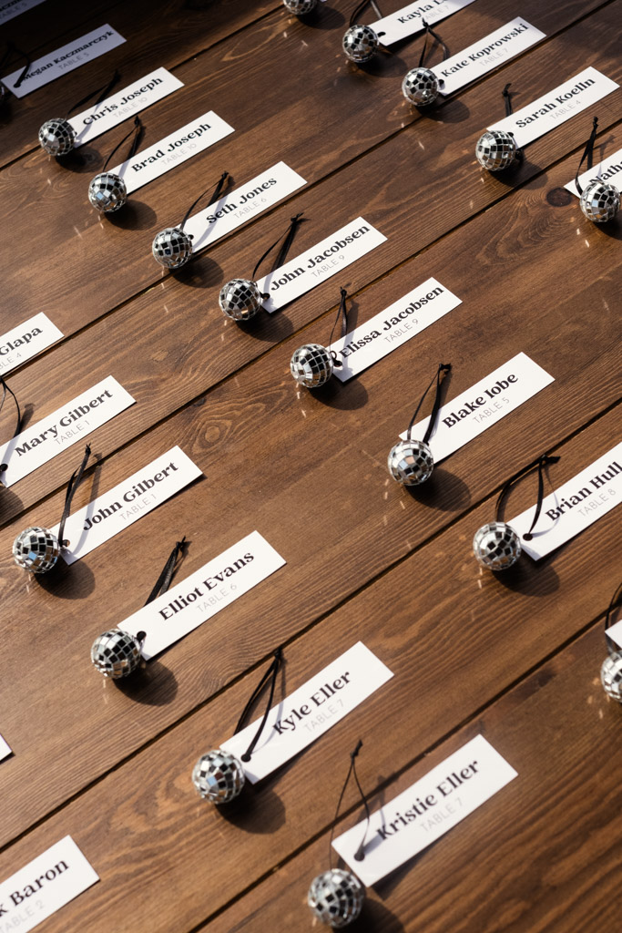 Name cards with small disco ball keychains are arranged on a wooden table in sunlight, for River Room wedding celebration