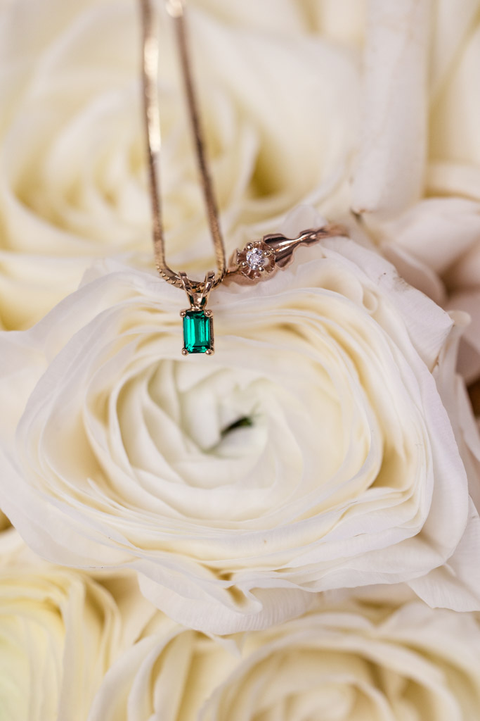 Gold necklace with a green gemstone and a diamond ring resting on white roses for River Room wedding celebration