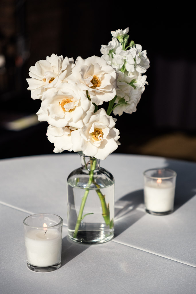White flowers in a glass vase with two lit candles rest on a white tablecloth, on a table for River Room wedding reception