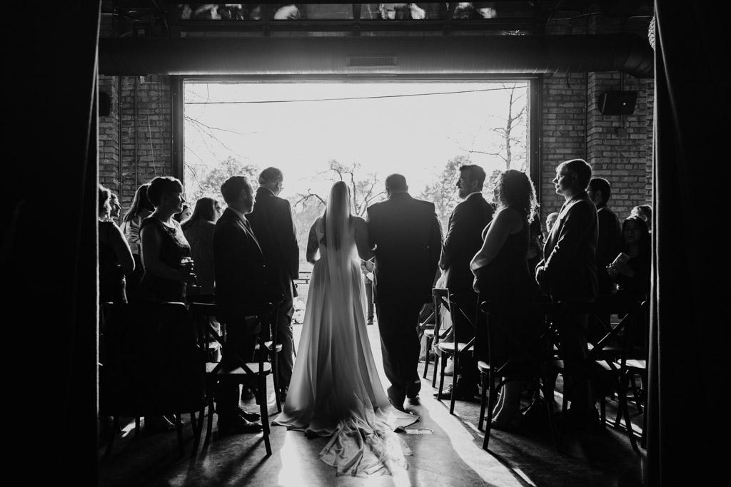 Bride walking down the aisle with her father in a sunlit River Room during wedding ceremony, guests watching