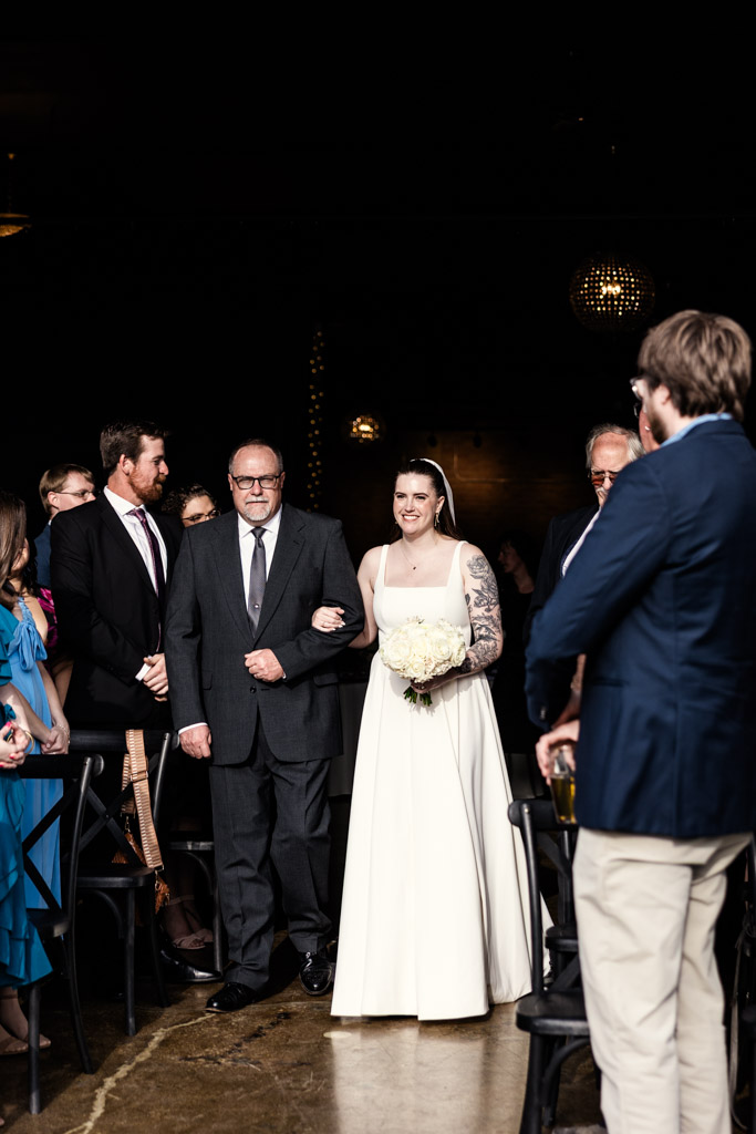 A bride in a white dress walks down the aisle with her father, surrounded by smiling guests during her River Room wedding ceremony