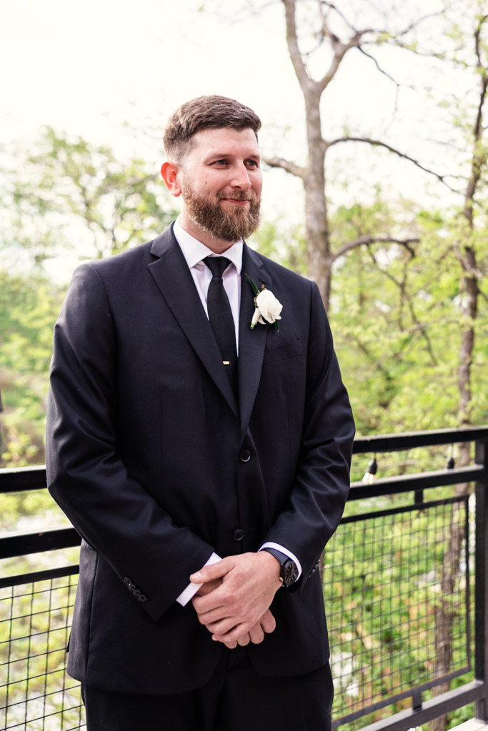 Groom in black suit with white rose boutonniere stands outside by a railing at River Room, with trees in the background
