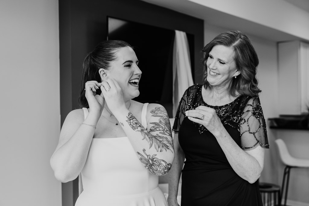 Bride and her mother laugh together as they get ready, the bride fastening earrings