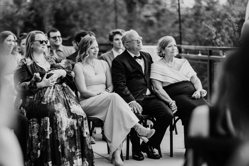 Groom's family sits in the front row at an outdoor River Room wedding ceremony, one holding a baby
