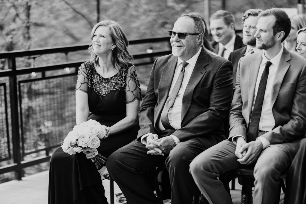 Bride's parents sit, smiling, during wedding ceremony at River Room. Other people are visible behind them