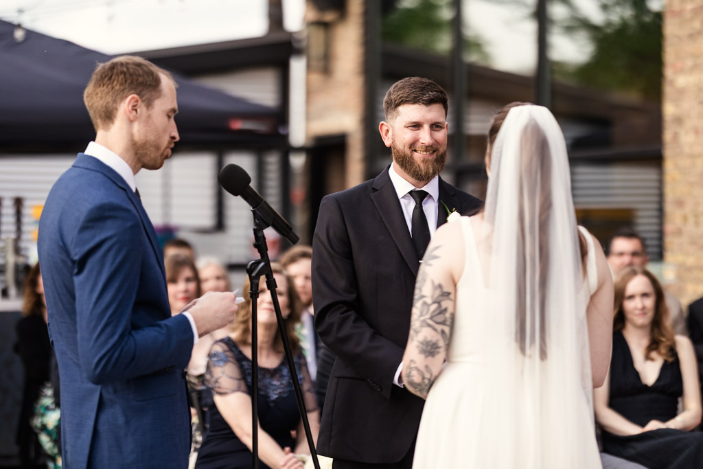 A groom smiles at his bride during an outdoor wedding ceremony at River Room, with guests seated behind them