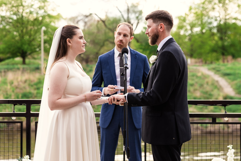 Bride and groom exchange rings outdoors at River Room, officiant in blue suit stands behind them