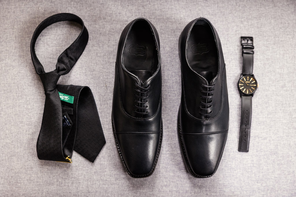 Groom's black dress shoes, a black necktie, and a black wristwatch arranged on a gray surface