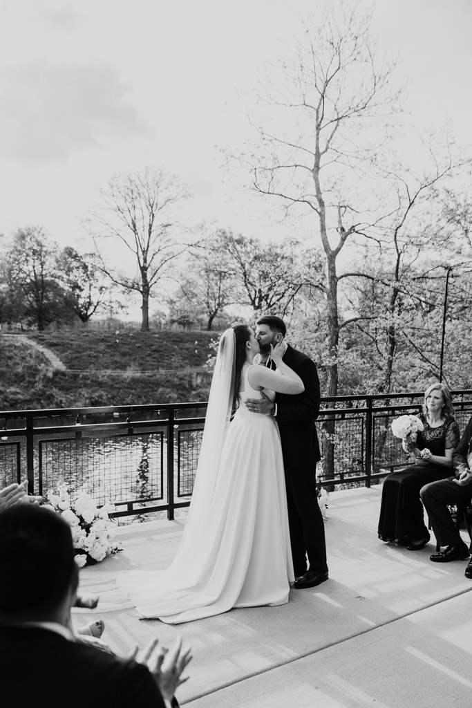 Bride and groom share their first kiss during their outdoor wedding ceremony at River Room, surrounded by guests