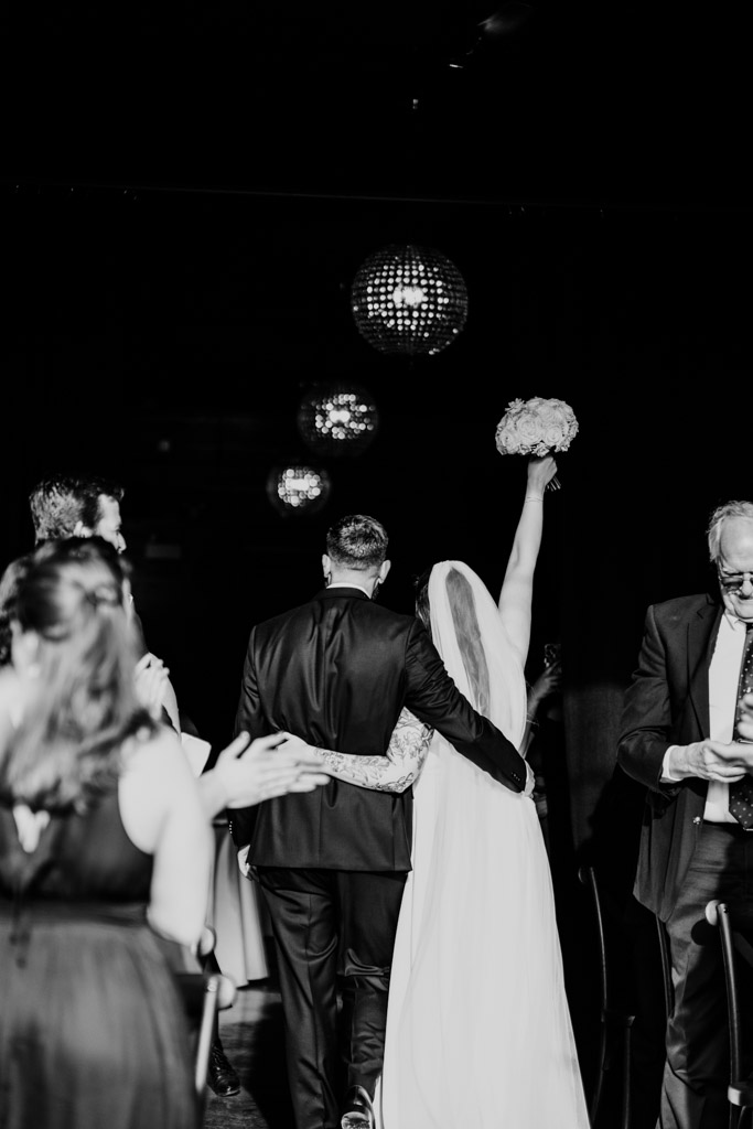 Bride and groom down the aisle arm-in-arm after their River Room wedding ceremony, the bride holds up her bouquet