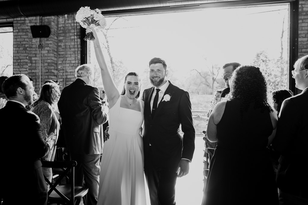 Black and white photo of happy bride raising her bouquet beside the groom inside River Room as guests look on after their wedding ceremony