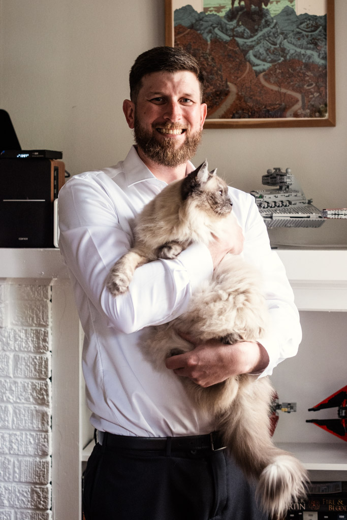 Groom in a white shirt smiling and holding a large fluffy cat while getting ready for his River Room wedding