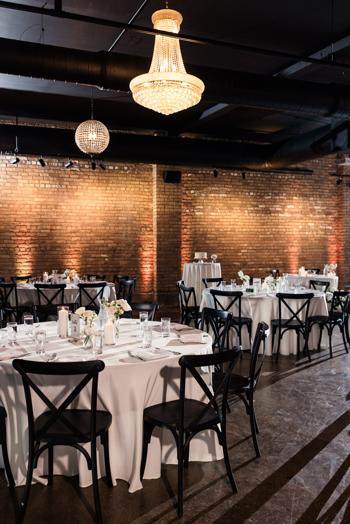 Chandelier-lit reception space with round tables, white tablecloths, black chairs, and brick walls at River Room