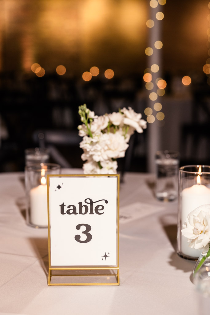 Table sign on a white-clothed table with candles and white flowers inside River Room at Burning Bush Brewery